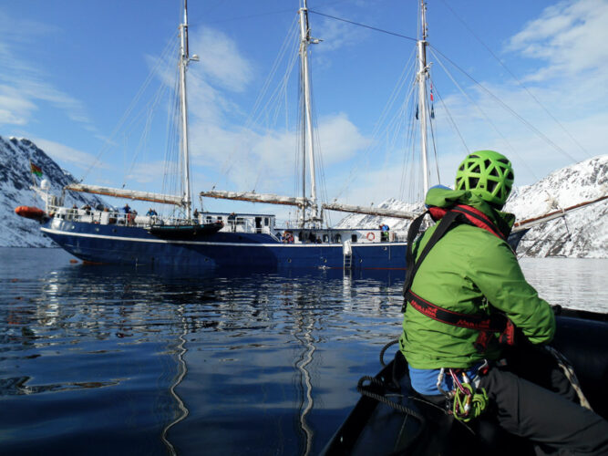 Person mit grüner Jacke auf einem Boot, fährt in Richtung größerem Boot, wenige Wolken am Himmel