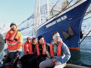 Einige Personen mit Rettungswesten blicken nach rechts und sitzen auf einem Schlauchboot, im Hintergrund ein großes blaues Boot