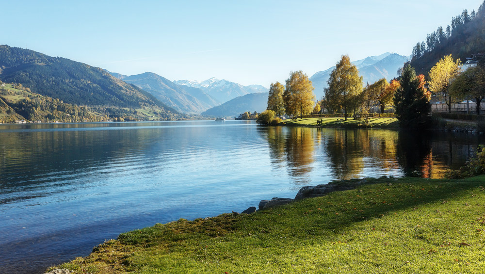 see mit klarem wasser an grasbewachsenem ufer, bunte bäume, berge im hintergrund