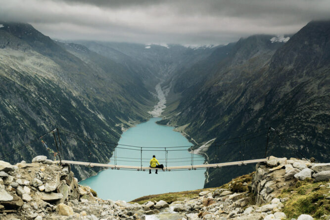 wanderer auf hängebrücke mit blick über hellblauen see, dunkle bergwände links und rechts