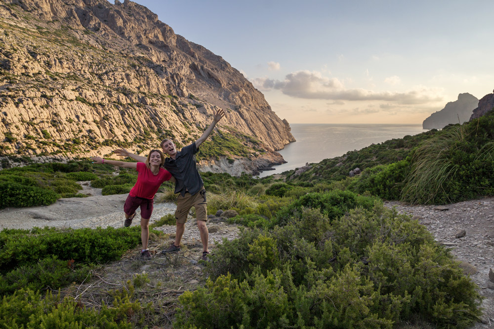 mädchen und junge freuen sich und strecken arme in die höhe, vor felsen und meer im hintergrund