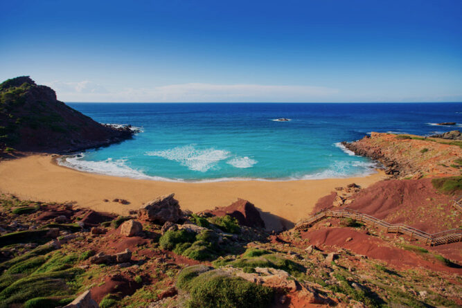 Bucht mit rötlichem Sand und hellblauem Wasser. Rote Hügel