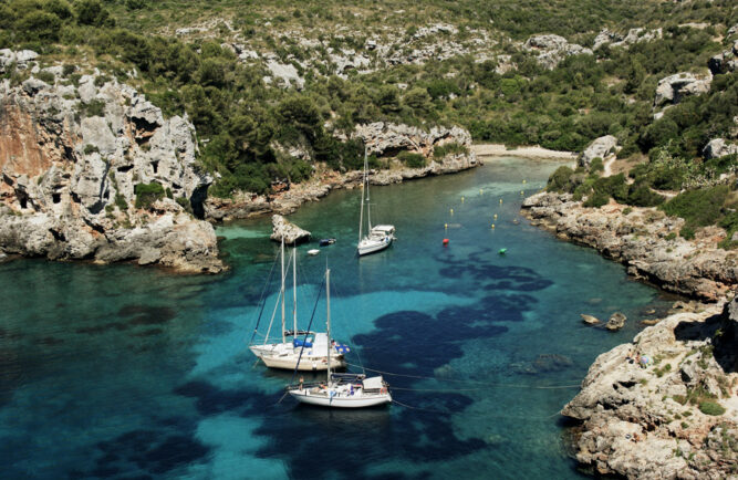 Weiße Segelboote auf seichtem, türkisem Wasser in einer Bucht. Felsige Wände und Wälder auf dem Festland