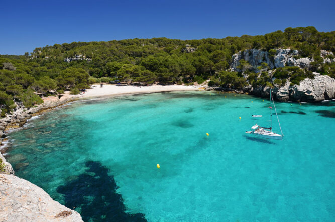 Eine Bucht mit einem weißen Sandstrand, hell türkisem Wasser. Ein Segelboot liegt in der Bucht. Hinter dem Strand sind grüne Wälder