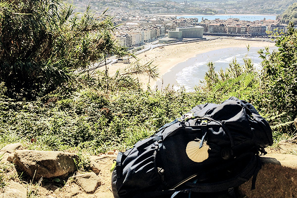 blauer Rucksack mit Wandermuschel über dem Strand von San Sebastián auf dem Camino del Norte in Spanien
