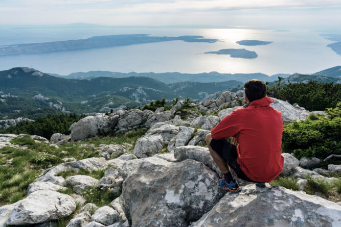 Mann mit roter Jacke sitzt auf Felsen über einer Küste