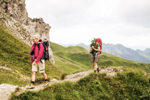 Zwei junge Frauen wandern entlang einem schmalen Wanderweg inmitten grüner Berglandschaft